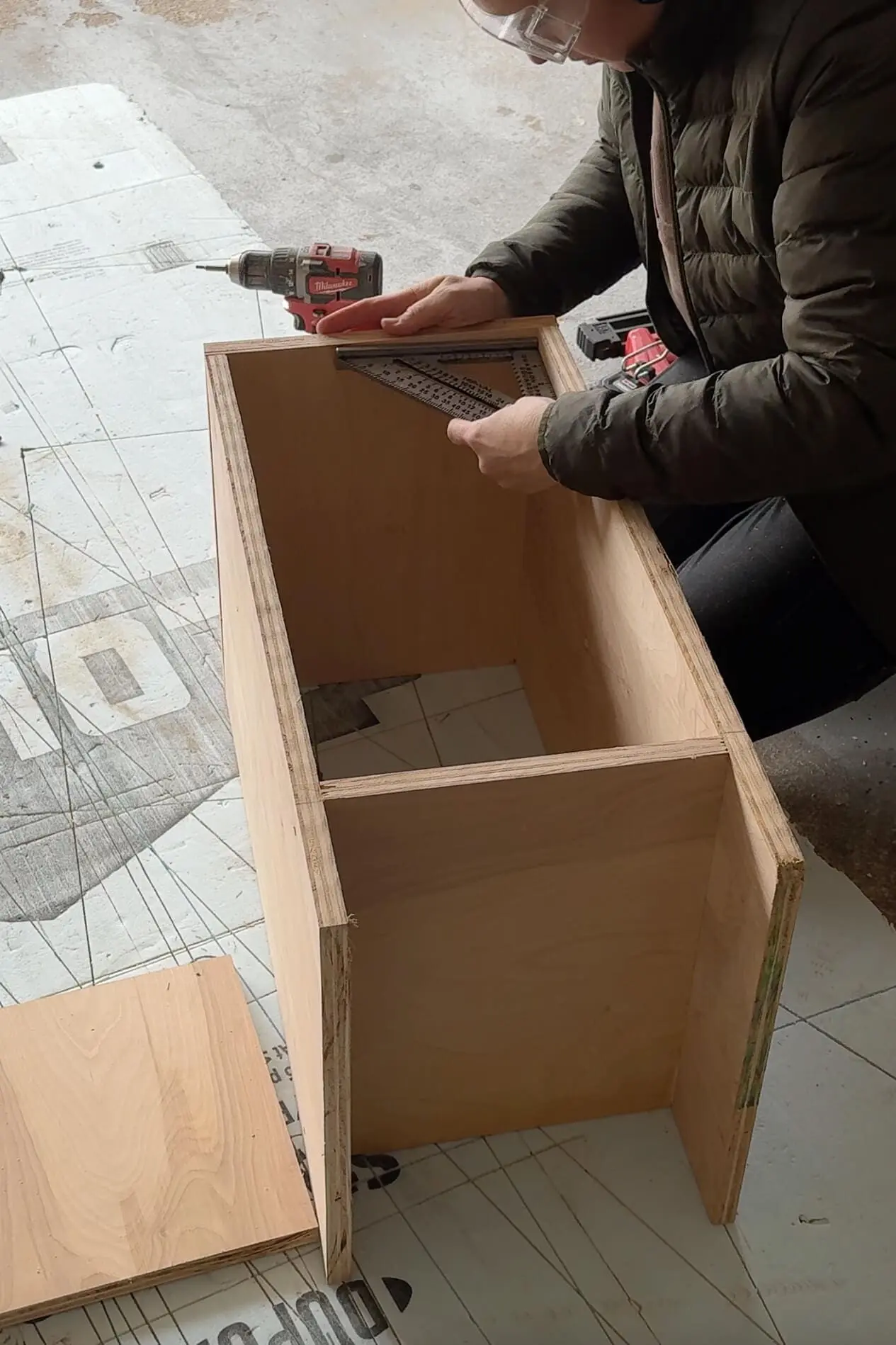 A DIYer measuring and checking the right angles of bedroom built ins using a metal square. She is wearing a puffer jacket and safety glasses, carefully ensuring the project is properly aligned before moving to the next step.